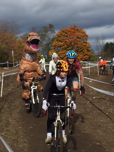 A group of costumed cyclists on a cyclocross course, with a T.Rex on the left.