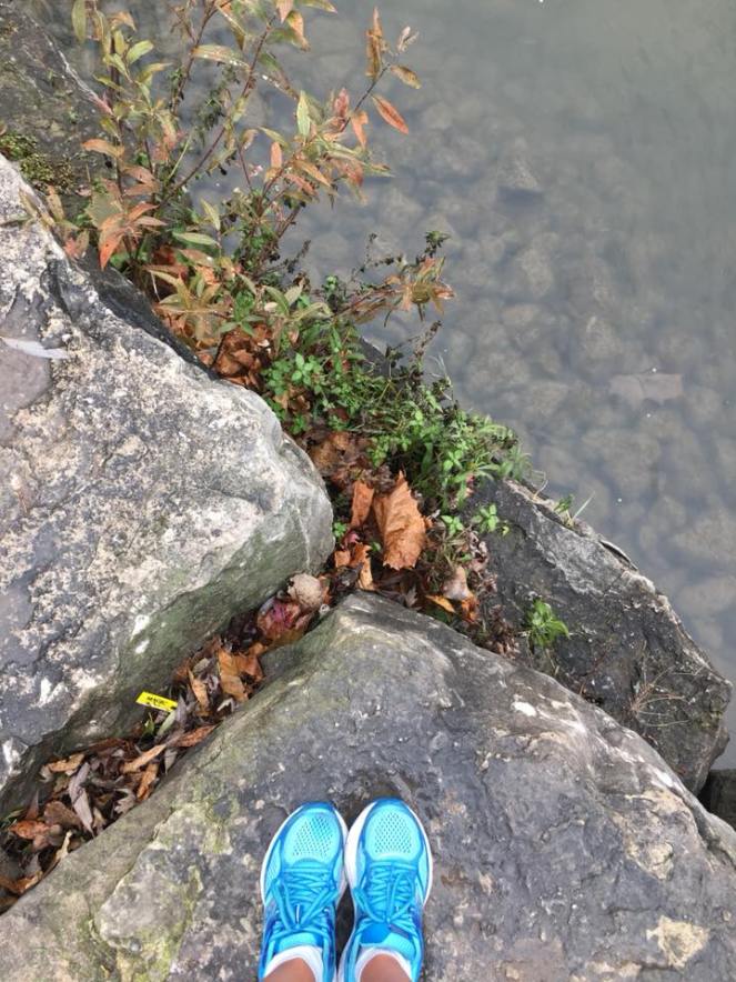 Image description: Tracy's two feet in robin blue running shoes on a stone slab beside the river, some dry leaves and greenery on the ground, the river cuts diagonally across the frame and you can see stones on the bottom through the clear water.