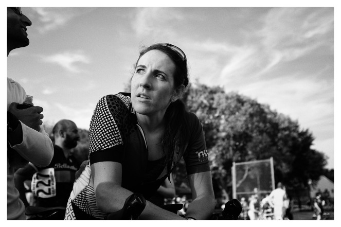  Erin, a dark haired woman with her hair pulled back, looks onto another spectator while wearing her cycling kit after one of her first cyclocross races. She is leaning forward on her bike. Her sunglasses are resting on top of her head, her jersey is zipped down, and her hair is wet from sweat. Photo Credit: Carlos Sabillon