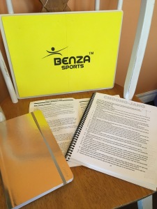 The author's tools for preparing for her test - a yellow rectangular plastic board for kicking, a gold notebook for recording her progress, and two white books printed with black type that include training theory and lists of patterns.