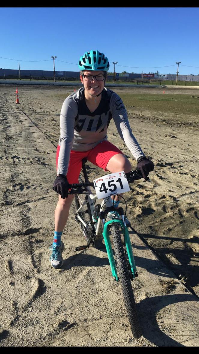 A 27-year-old woman wearing a grey shirt and pink shorts, sitting on her bike on a dirt track, smiling at the camera.