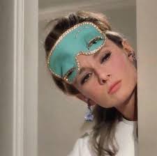 Actress Audrey Hepburn as Holly Golightly in Breakfast at Tiffany's. She is a slender, brown-haired woman and she looking out around her apartment door. Only her head and one shoulder can be seen, she is wearing a light blue sleep mask pushed up on her forehead and there is a tassel visible, dangling from her right ear. 