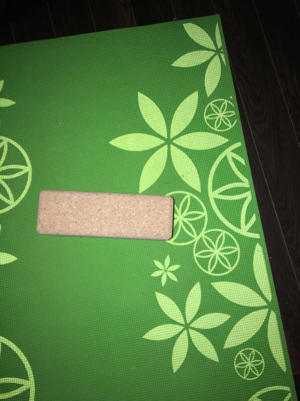 A rectangular, light brown cork yoga block sits atop a green yoga mat that is decorated with lighter green flowers. Dark brown floorboards are in the background.