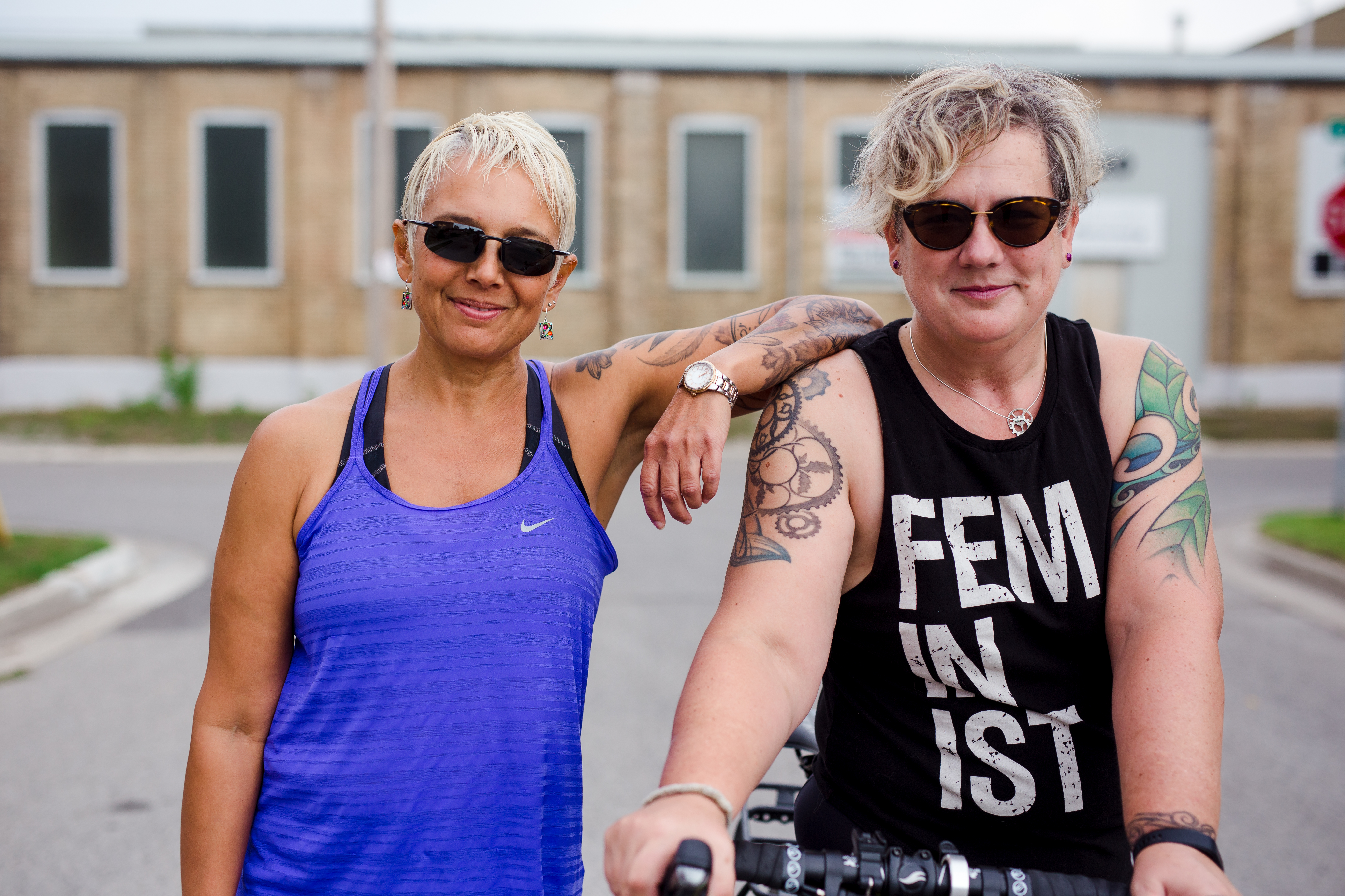 Image description: Street shot of Tracy (left) and Sam (right) from the waist up. Tracy is in a running tank, leaning on Sam's shoulder with her left elbow; Sam in on her bike, wearing a tank that says FEMINIST. Both are smiling, wearing sunglasses, street and building in background.