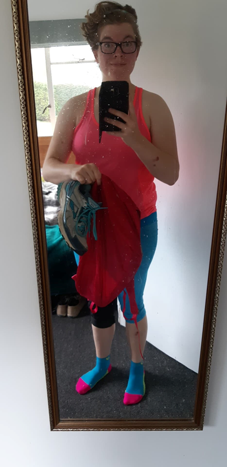 A 29-year-old white woman taking a selfie in a mirror. She is wearing a pink tank top, blue knee-length leggings, a black knee brace, and turquoise, berry, and lime socks. She is holding a berry-coloured backpack and a pair of grey and turquoise runners.