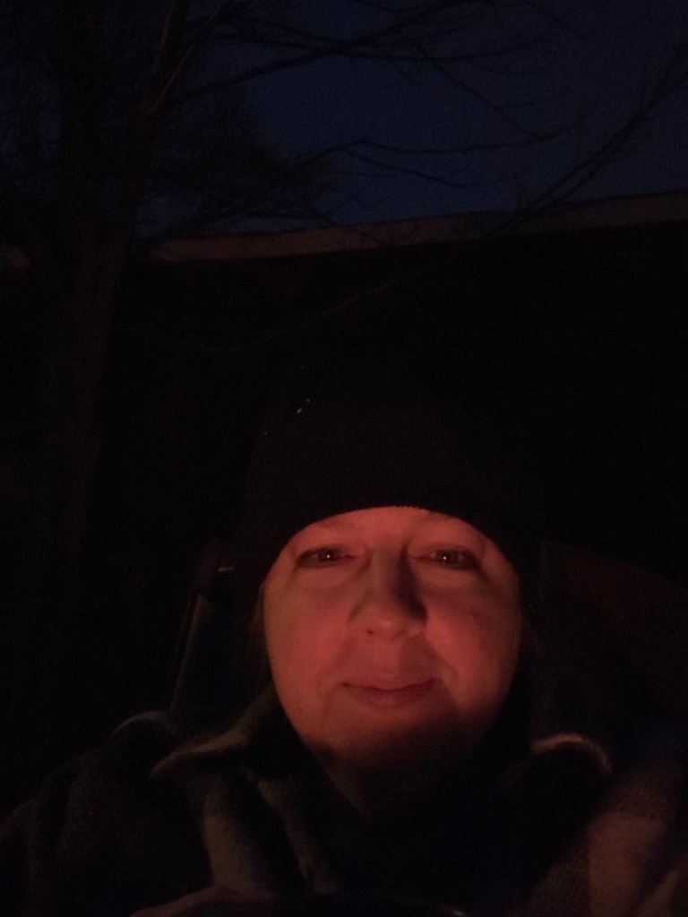 A nighttime selfie of a woman with a round face wearing a dark hat and dark clothes, she is lit by firelight.