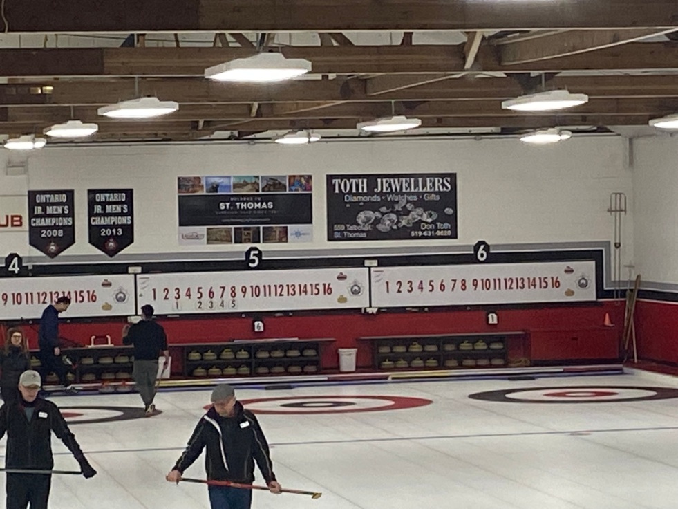 An image of curling sheets of ice with players on the ice watching the curling rock. Scoreboards on the back wall.