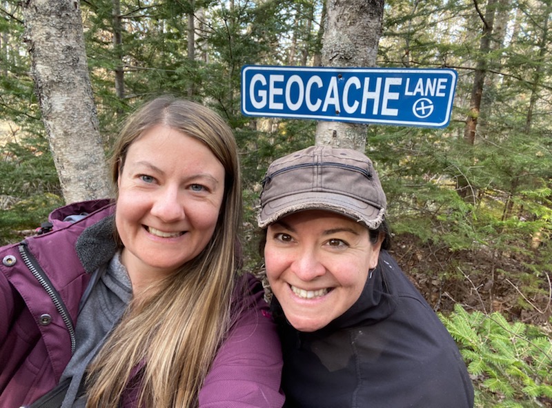 Alispice and Elan at the oldest geocache in Canada. Trees and a sign that says Geocache Lane behind them.