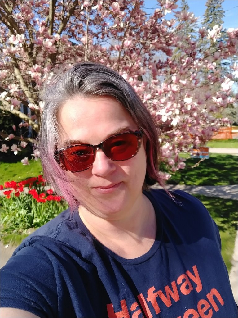 Selfie of a woman with greying hair and brown sunglasses in front of a blooming pink magnolia tree. She has bright sunlight on her face and is wearing a navy coloured tshirt reading "halfway between"